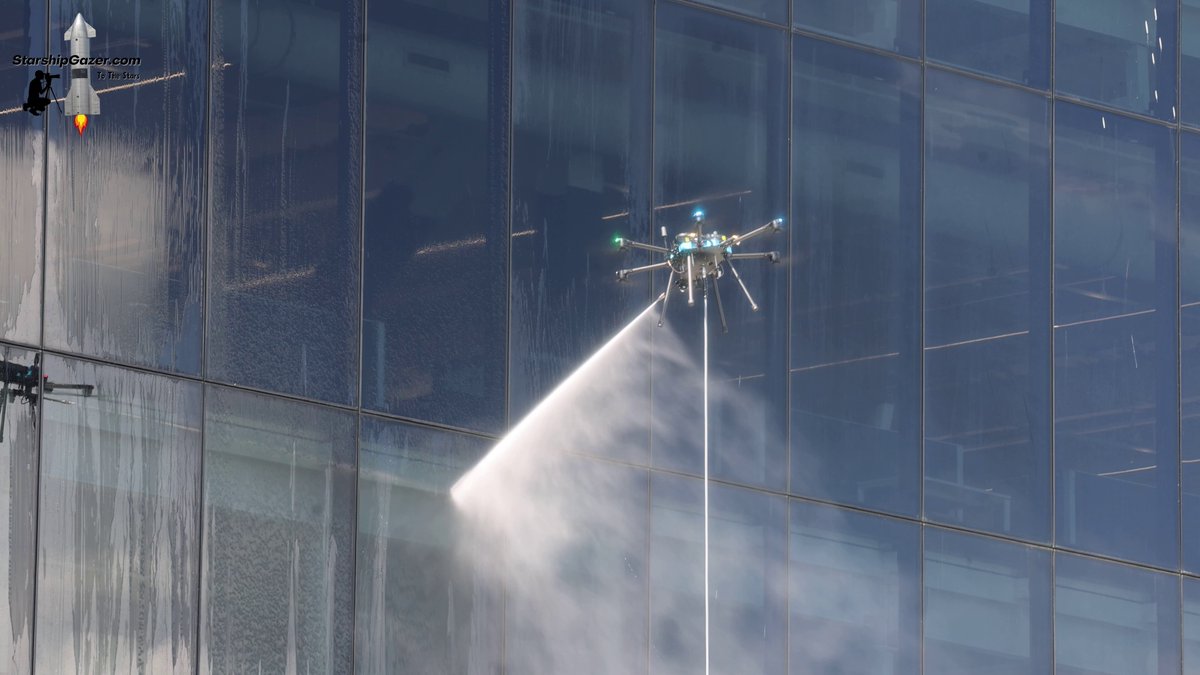 A large multirotor drone sprays water across the glass facade of Starbase's launch control room.