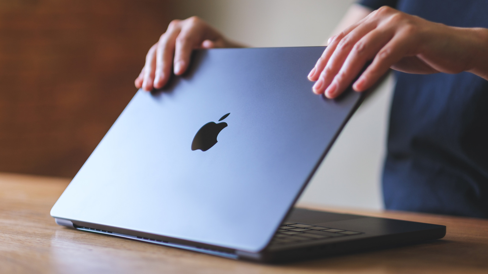 A person closes a silver MacBook Pro on a wooden table.