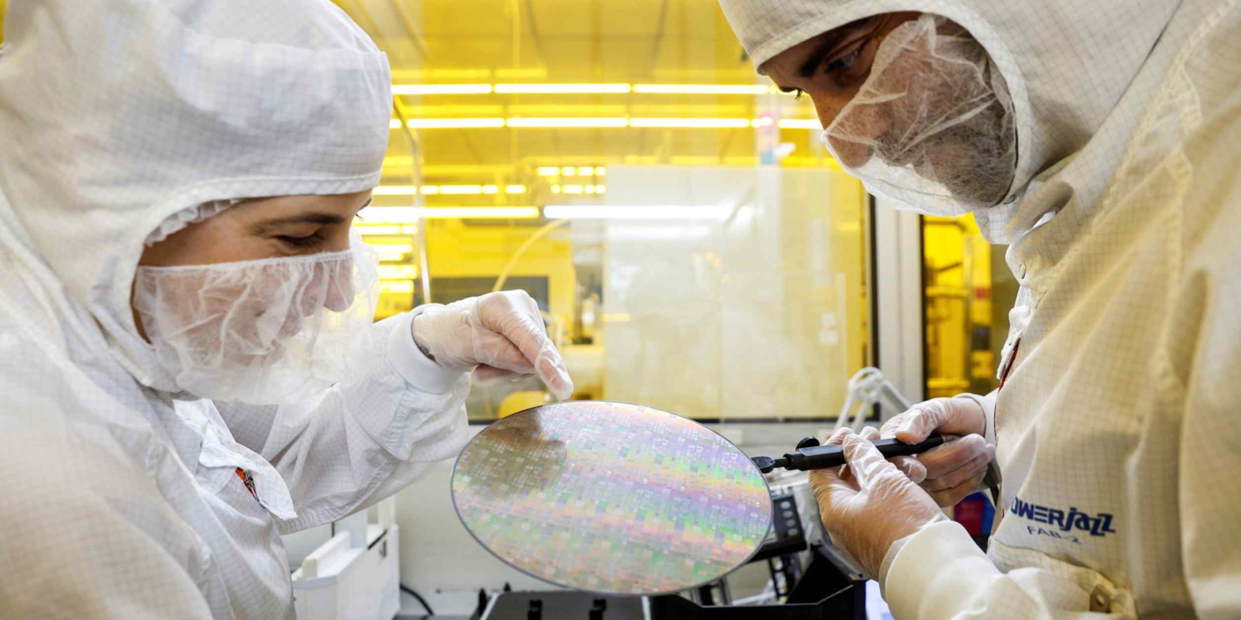 Two clean-room workers inspect a semiconductor wafer at a Tower Semiconductor facility.